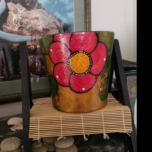 Puede incluir: Una maceta pintada a mano con un gran diseño de flor roja. La flor tiene un centro amarillo con puntos blancos y negros. La maceta está sobre una esterilla de bambú y un soporte negro. El fondo incluye una imagen enmarcada y conchas marinas.