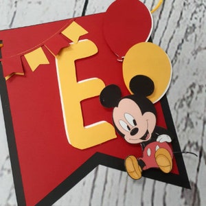 Pu&ograve; includere: Un banner rosso con bordo nero, con la lettera gialla "E" con contorno bianco, un cartone animato di Topolino e palloncini rossi e gialli. &Egrave; inclusa anche una ghirlanda di bandierine gialle e rosse.