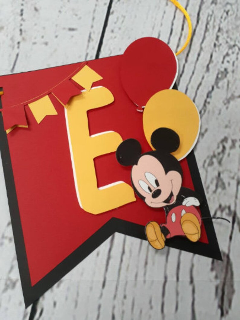 Pu&ograve; includere: Un banner rosso con bordo nero, con la lettera gialla "E" con contorno bianco, un cartone animato di Topolino e palloncini rossi e gialli. &Egrave; inclusa anche una ghirlanda di bandierine gialle e rosse.