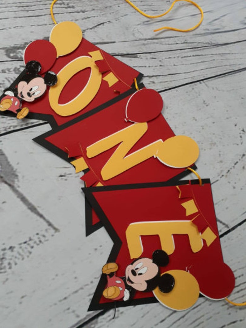 Pu&ograve; includere: Un banner rosso, nero e giallo con la scritta "ONE" in lettere gialle. Il banner presenta ritagli di Topolino e palloncini rossi e gialli. Il banner &egrave; infilato su una corda gialla.