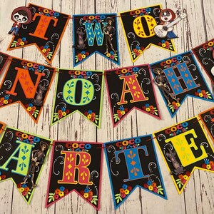 May include: A colorful banner set featuring characters from the movie Coco. The banner spells out "TWO" and "CARTER NOAH" in vibrant letters. Each pennant is black with colorful floral accents and character cutouts, perfect for a birthday celebration.