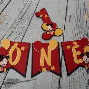 Pu&ograve; includere: Un banner rosso, nero e giallo con la scritta "ONE" in lettere gialle. Il banner presenta ritagli di Topolino e palloncini. &Egrave; presente anche un ritaglio separato del numero "1" con Topolino. Il banner &egrave; infilato con uno spago giallo.