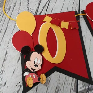 Pu&ograve; includere: Un banner rosso e nero con il numero giallo "0" e Topolino. Il banner presenta anche palloncini gialli e rossi e piccole bandierine rosse. Lo striscione &egrave; infilato su un cordino giallo.