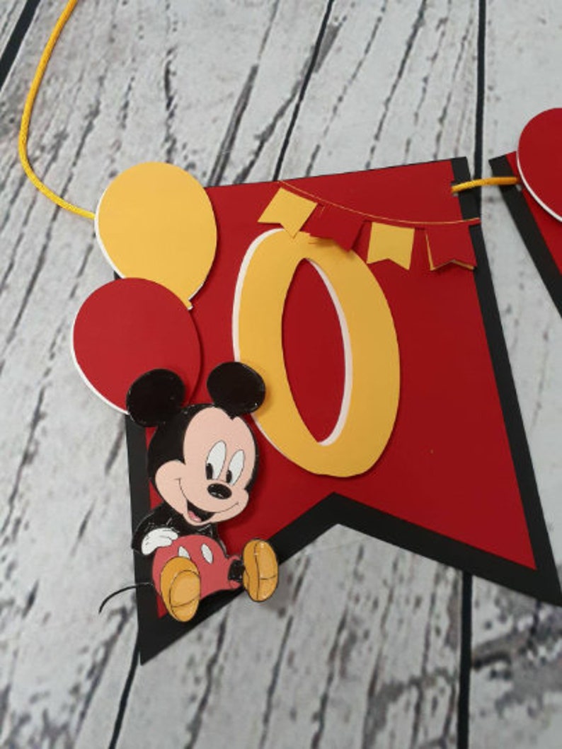 Pu&ograve; includere: Un banner rosso e nero con il numero giallo "0" e Topolino. Il banner presenta anche palloncini gialli e rossi e piccole bandierine rosse. Lo striscione &egrave; infilato su un cordino giallo.