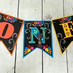 May include: A vibrant banner spelling "ONE" with characters from the film Coco. Each flag is black with a different coloured border and character. The banner is strung on twine, ideal for a birthday.