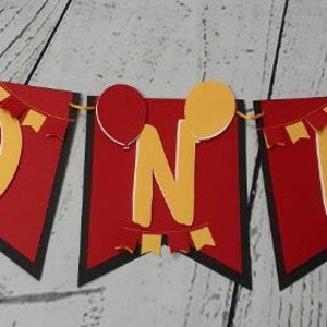 Pu&ograve; includere: Un banner rosso, nero e giallo che dice "ONE" con motivi di Topolino e palloncini. Il banner &egrave; infilato su un cordino giallo, perfetto per una festa di primo compleanno.