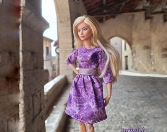 Puede incluir: Una muñeca de pelo largo rubio viste un vestido morado con estampado moteado y una cintura plateada brillante. El vestido tiene mangas tres cuartos y falda acampanada. El texto AnnaGì design es visible en la esquina.
