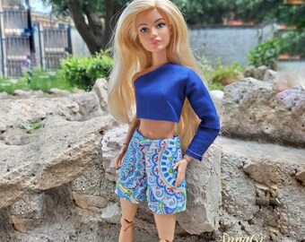 Puede incluir: Una muñeca rubia viste un top corto de una sola manga larga en color azul real y pantalones cortos estampados con un diseño colorido estilo mandala en tonos azules, verdes y morados. La muñeca tiene articulaciones y cabello rubio largo.