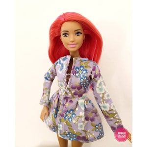 Può includere: Una bambola che indossa un vestito viola con una stampa floreale. Il vestito ha maniche lunghe e un collo a V. La bambola ha capelli lunghi e rosa brillante.