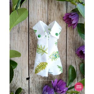 Può includere: Un vestito di stoffa bianca per bambole con una stampa di foglie verdi e bottoni verdi. Il vestito è piegato ed esposto su una superficie di legno con foglie verdi e fiori viola.