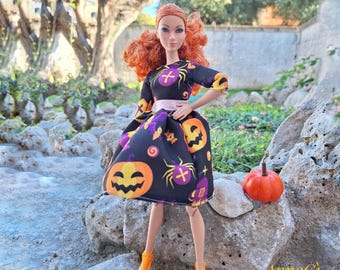 Puede incluir: Una muñeca con cabello naranja vibrante viste un vestido negro con un patrón de calabazas de Halloween naranjas, arañas moradas y cruces. El vestido tiene una cintura plateada brillante y mangas tres cuartos. La muñeca lleva zapatos naranjas.