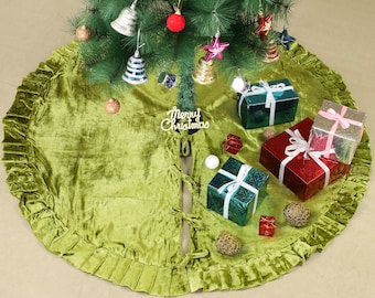 Puede incluir: Una falda de árbol de Navidad de terciopelo verde con un borde con volantes. La falda tiene una abertura en la parte posterior con dos lazos. La falda está decorada con regalos, adornos y un letrero que dice "Feliz Navidad".