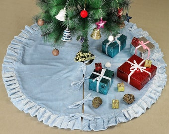 Puede incluir: Una falda de árbol de Navidad de terciopelo azul claro con un borde con volantes. La falda está decorada con lazos dorados y un letrero que dice "Feliz Navidad". Hay varios regalos envueltos y adornos en la falda.