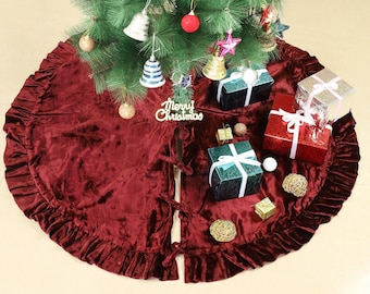 Puede incluir: Una falda para árbol de Navidad de terciopelo rojo intenso con un borde con volantes y una abertura central con cierres de lazo. La tela tiene una textura suave y arrugada. Un letrero blanco con el texto Merry Christmas está en el centro.