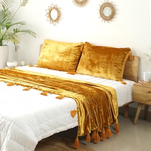 May include: A mustard yellow velvet throw blanket with tassels on a white bed with matching velvet pillows.