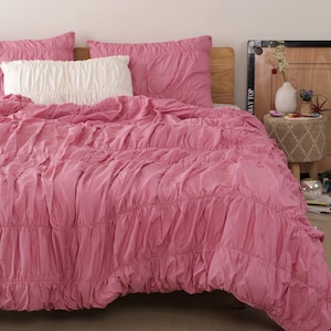 May include: A pink bedspread with a ruched design, paired with matching pink pillowcases and a white ruched pillow. The bedding is arranged on a wooden bed frame, creating a cozy and inviting bedroom setting.