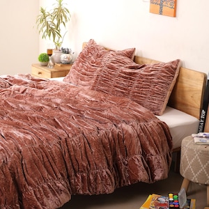 以下が含まれることがあります： ダスティローズのベルベットの掛け布団と、お揃いの枕カバー2つでベッドが作られています。寝具はギャザーの質感とフリルの縁取りが特徴です。ベッドフレームは明るい色の木材で作られています。