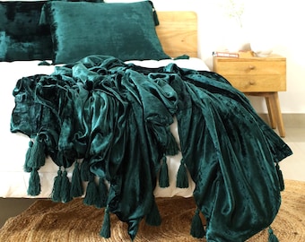 Puede incluir: Una manta de terciopelo verde oscuro con borlas en los bordes. La manta está drapeada sobre una cama con almohadas de terciopelo verde a juego.