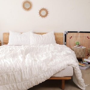 May include: White ruffled duvet cover and two matching pillowcases on a bed with a wooden headboard. The bed is made with a white comforter and the bedding is arranged in a simple and elegant style.