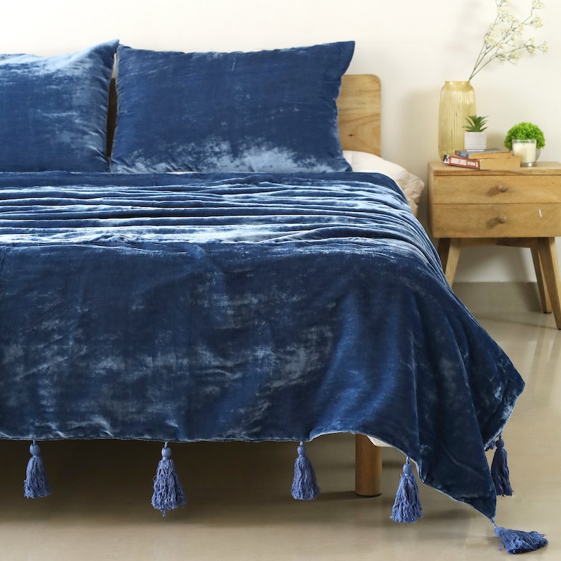 May include: A blue velvet throw blanket with tassels on the edges. The blanket is draped over a bed with blue velvet pillows.