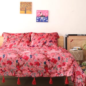 May include: Pink floral bedspread and pillow set featuring a bird and floral design. The bedspread has red tassels along the bottom edge. Two small paintings hang on the wall above the bed.