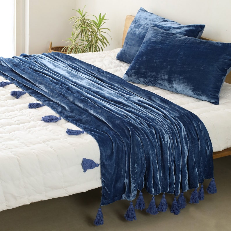 May include: A blue velvet throw blanket with tassels on a white bed. The blanket is folded and draped over the bed, with the tassels hanging down the sides.