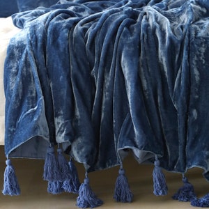 May include: A soft, blue velvet throw blanket with a subtle shimmer and tassel trim. The blanket is folded and draped over a bed.