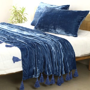 May include: A blue velvet throw blanket with tassels on a white bed. The blanket is folded and draped over the bed with the tassels hanging down the side.