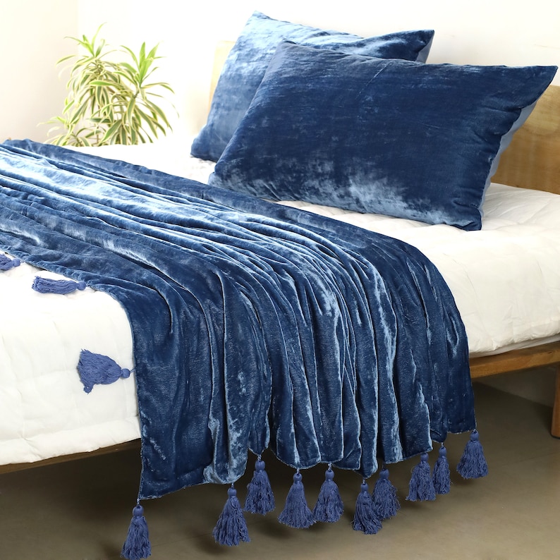 May include: A blue velvet throw blanket with tassels on a white bed. The blanket is folded and draped over the bed with the tassels hanging down the side.