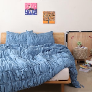 May include: A light blue bedspread and pillow set with a gathered, textured design. The bedding is on a wooden bed frame. Two small paintings hang on the white wall above the bed.