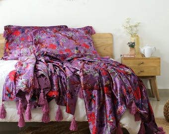 May include: A bed set featuring a purple velvet throw blanket and two pillowcases. The bedding has a floral pattern in red, green, and purple. The throw blanket has purple tassels along the edges. The bed is made up with white sheets.