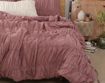 Puede incluir: Una cama con un edredón de color rosa polvoriento y fundas de almohada a juego. También hay una almohada blanca en la cama. El edredón tiene un diseño fruncido. La cama está hecha y lista para usar.