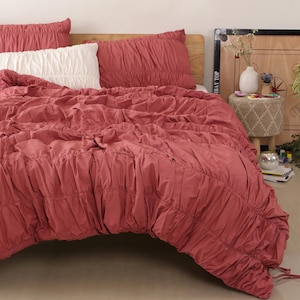 Peut inclure: Un lit est fait avec une housse de couette rose poussiéreux et des taies d'oreiller assorties. Un oreiller blanc est également sur le lit. La housse de couette a un design ruché. Le lit est contre une tête de lit en bois.