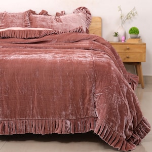 May include: A dusty rose velvet bedspread and matching pillow shams featuring a ruffled edge trim. The fabric has a soft, crushed texture with a subtle sheen. The bedding set covers the mattress and drapes over the sides of the bed frame.