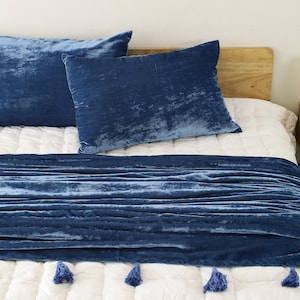May include: A blue velvet throw blanket with tassels on a white bed with blue velvet pillows.
