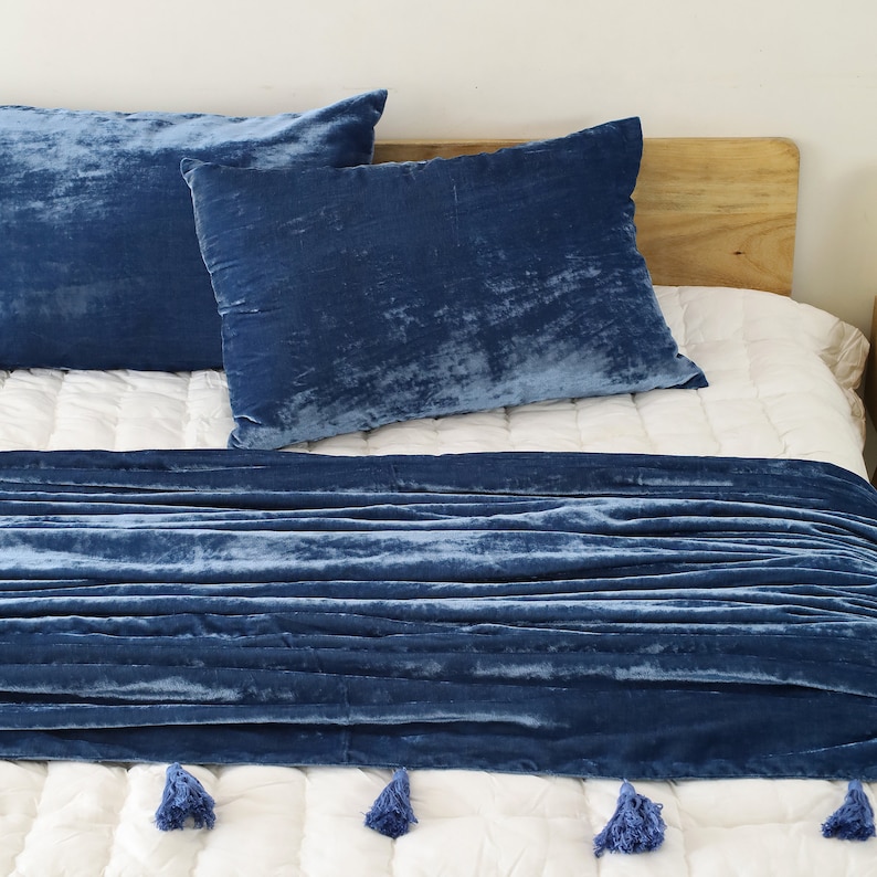 May include: A blue velvet throw blanket with tassels on a white bed with blue velvet pillows.
