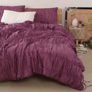 May include: A bed is made with a plum-coloured comforter and matching pillowcases. A white pillow is placed in front of the plum-coloured pillows. The comforter has a ruched design. The bed is set against a wooden headboard.