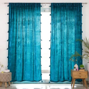 May include: A pair of floor-length teal velvet curtains hanging from a silver metal rod. The curtains feature a soft, reflective texture and are adorned with small, matching teal tassels attached along the outer vertical edges of each panel.