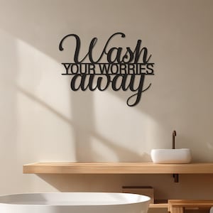 May include: Black metal wall art with the words "Wash Your Worries Away" in a cursive font. The sign is mounted on a light beige wall above a wooden shelf with a white sink and tap.