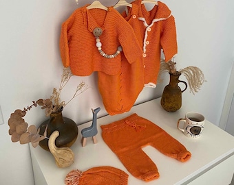 Könnte beinhalten: Ein Set orangefarbener gestrickter Babykleidung, bestehend aus einem Cardigan, einem Strampler, einer Hose und einer Mütze. Die Kleidung liegt auf einer weißen Oberfläche.