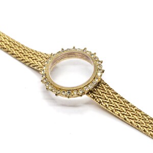 May include: A gold-tone watch with a round face and a clear crystal bezel. The watch has a gold-tone metal band with a woven design.