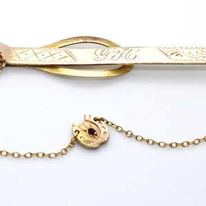 Op de afbeelding: Een gouden stropdasgesp met een ketting die is bevestigd aan een hoefijzerbedel. De stropdasgesp heeft een gegraveerd ontwerp en de initialen "Y.H." aan de voorkant.