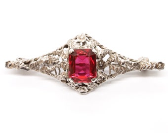May include: A silver filigree brooch with a large, red, square-shaped gemstone.