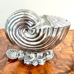 May include: A silver-coloured, nautilus-shaped decorative box with a hinged lid. The box sits on a detailed base with a seashell accent. The surface of the box has a spiral design, and the lid is decorated with an engraved pattern.