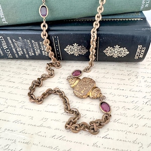 Peut inclure: Collier ancien de couleur or avec un pendentif décoratif et des pierres précieuses ovales violettes. Le collier est présenté sur un fond de livres anciens et de papier manuscrit, créant une esthétique classique et vintage.