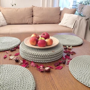 Könnte beinhalten: Ein Set aus sechs gehäkelten Platzsets in Salbeigrün auf einem Holztisch mit roten Herbstblättern und Kastanien, die verstreut liegen. Ein Stapel der Platzsets befindet sich in der Mitte des Tisches mit einer Holzschale mit roten Äpfeln darauf.