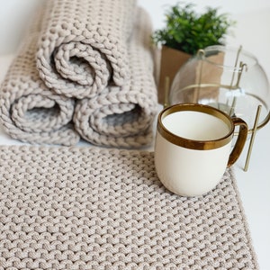 Könnte beinhalten: Ein beiger gestrickter Untersetzer mit einem braunen Lederanhänger mit der Aufschrift "Matt.va" liegt auf einer weißen Oberfläche. Eine weiße Tasse mit goldfarbenem Rand steht auf dem Untersetzer. Im Hintergrund liegt ein Stapel beiger gestrickter Rollen.