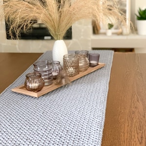 May include: A gray knitted table runner with a wooden tray holding a bouquet of dried pampas grass in a white vase and several clear glass candle holders.