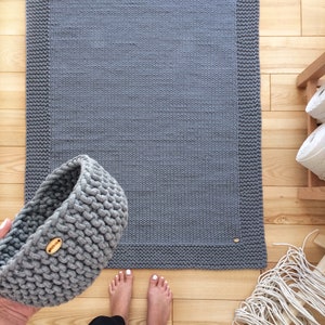 May include: A gray knitted rug with a thick border. The rug is on a wooden floor. A gray knitted basket is in the foreground.