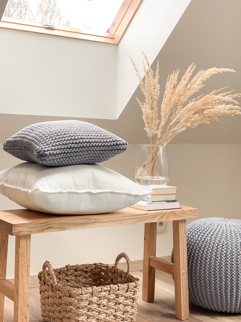 May include: A wooden bench with a gray knitted pouf, a woven basket, and two gray knitted pillows. A vase with dried pampas grass sits on the bench.